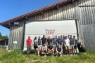 Menschen stehen vor einem Holzhaus. 